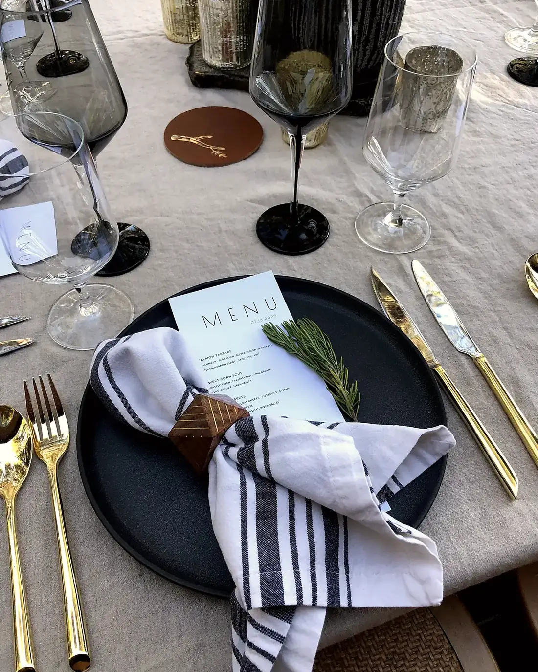 A single place setting on linen: a matte black charger, striped napkin in a wooden ring, gold flatware, crystal stemware and a printed menu card
